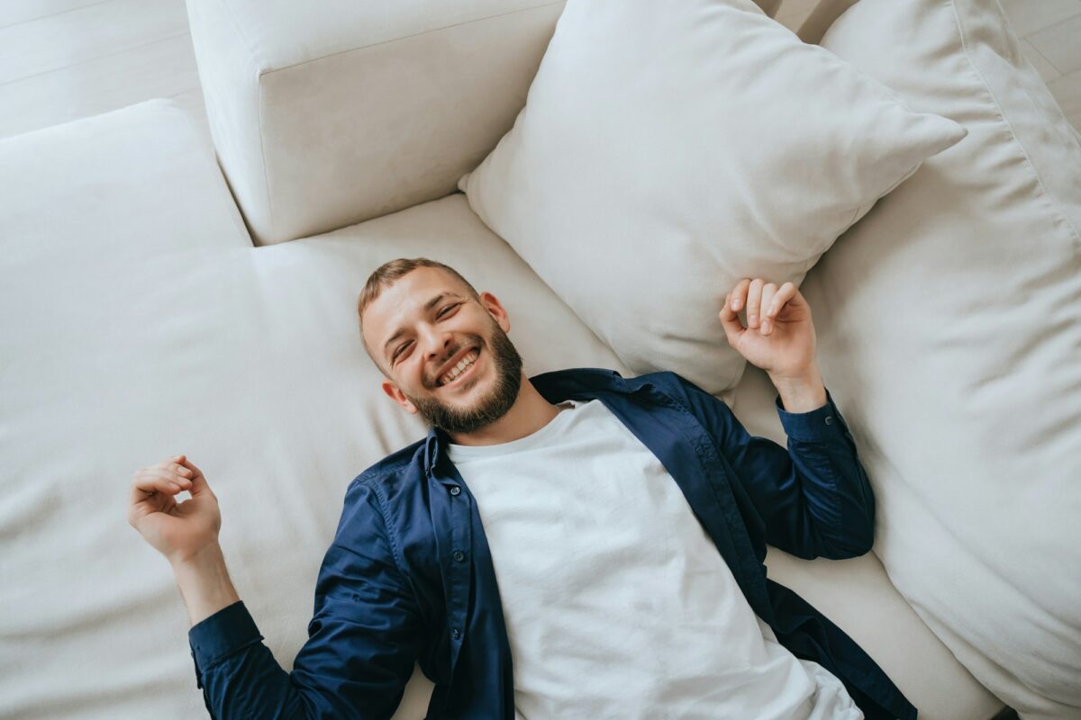 Homme heureux, barbu, en tenue décontractée, allongé sur un canapé avec des coussins, souriant, regardant la caméra, se relaxant à la maison.