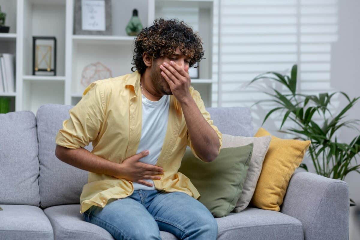 Jeune homme malade à la maison, assis sur le canapé, l'Américain d'origine latine souffre de fortes douleurs à l'estomac et de nausées, il a été empoisonné.