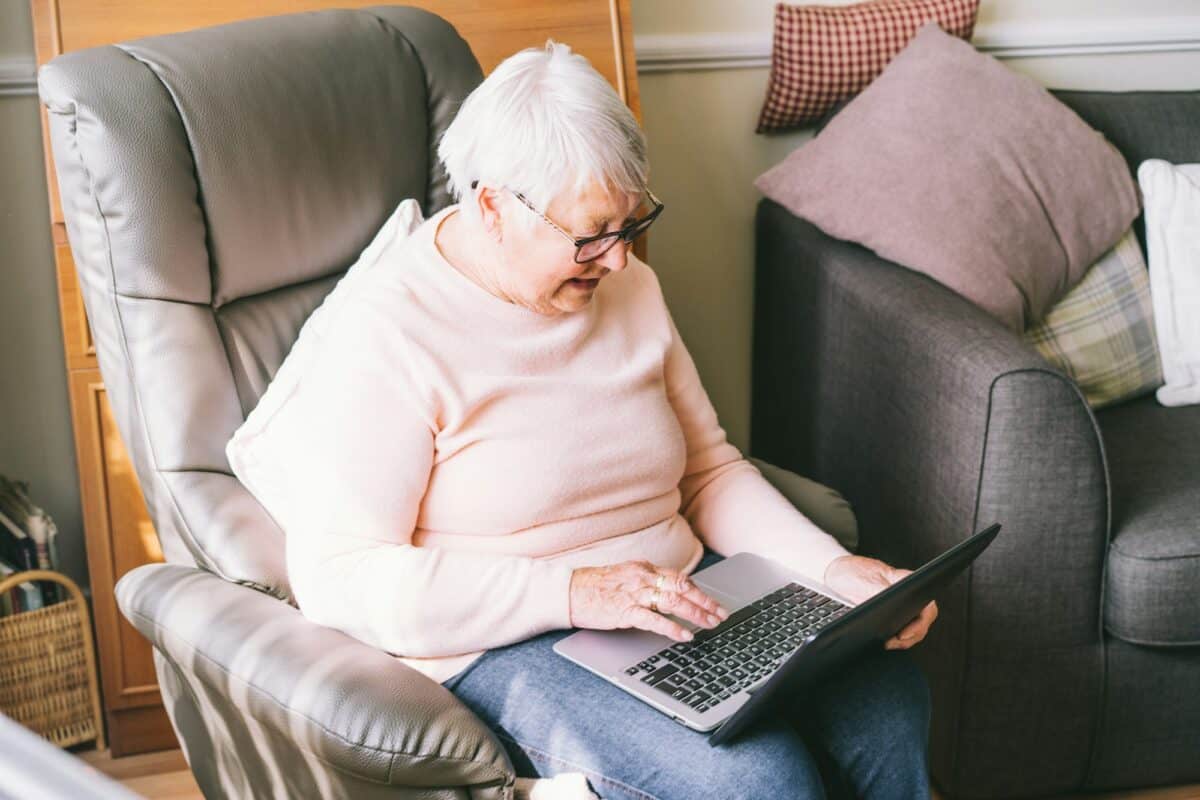 The hoary senior grandmother is sitting on the armchair, looking at the laptop screen.